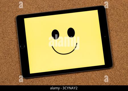 Nahaufnahme eines Tablets mit einem menschlichen lächelnden Gesicht, das Glücksgefühl auf gelbem Hintergrund und Korkhintergrund repräsentiert Stockfoto