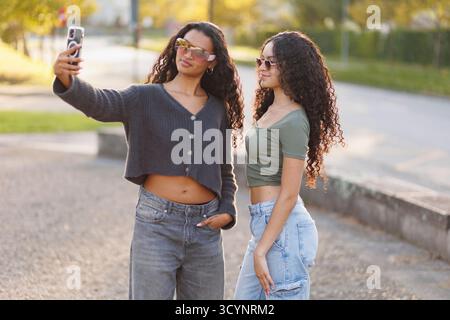 Zwei junge Erwachsene Frauen mit Sonnenbrille und lässiger Kleidung posieren während der Golden Hour für ein Selfie und halten einen Moment der Freundschaft und des modernen Lebensstils fest Stockfoto