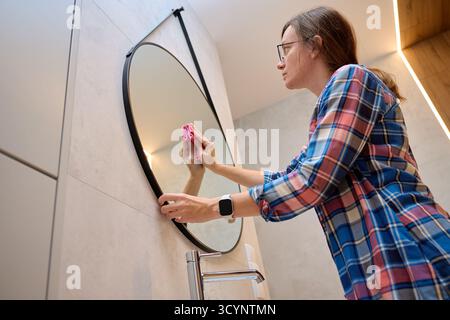 Frau mit Brille, die im modernen Badezimmer einen runden Spiegel mit rosafarbenem Tuch reinigt. Konzept der Hausreinigung, Hygiene, Alltag und Hausarbeit Stockfoto