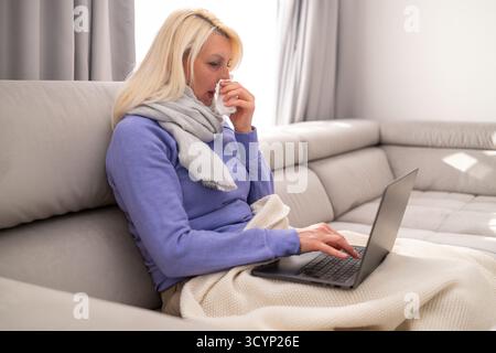 Blonde Frau mittleren Alters, die mit einer Decke auf der Couch sitzt, sich unwohl fühlt und zu Hause Filme auf ihrem Laptop ansieht. Konzept von Krankheit, Ruhe, Entspannung und gemütlicher häuslicher Atmosphäre. Stockfoto