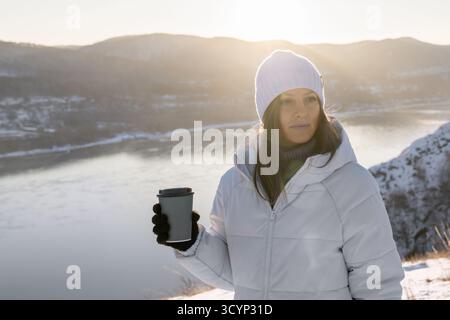 Eine schöne Frau im Winter in einer warmen, gemütlichen weißen Jacke bewundert die Landschaft mit einem Wald und einem Fluss während des Kaffees. Nur Kontemplation und Stockfoto