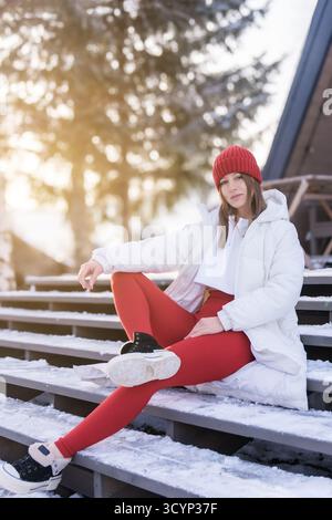 Junge Frau in Wintermode, die sich bei Tageslicht auf verschneiten Stufen in sonniger Umgebung im Freien entspannt Stockfoto