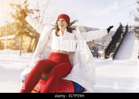 Eine junge Frau mit einem hellen Lächeln genießt den Nervenkitzel, einen schneebedeckten Hang auf einem aufblasbaren Schlauch hinunter zu gleiten. Sie ist warm in einer weißen Jacke gekleidet Stockfoto