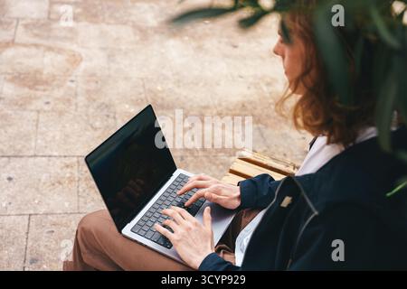 Junge Person, die an einem Laptop arbeitet und auf einer Bank im Freien sitzt Stockfoto