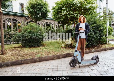Junge Person, die auf einem Bürgersteig neben einem elektrischen Roller steht und ein Smartphone benutzt Stockfoto