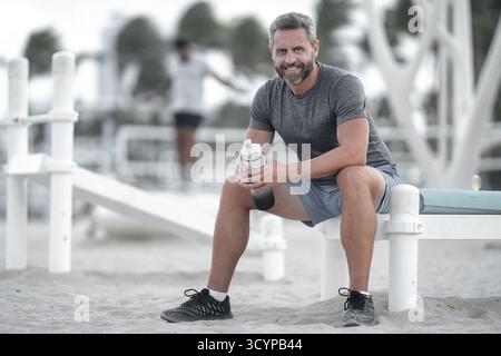 Sport-Fitness-Mann, der draußen auf einer Bank ruht. Reifes Fitnesssportmodell, das nach dem Training auf einer Bank sitzt. Ein Sportmann macht nach dem Fitnesstraining eine Pause Stockfoto
