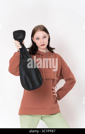 Junge Ukulele-Spielerin, die Ukulele in der Hand hält und den Arm nach vorne streckt. Die 18-jährige kaukasische Musikerin trägt einen braunen Kapuzenpullover und ein Ligh Stockfoto