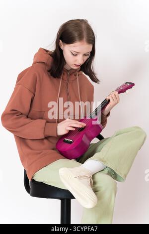 Ukulelistin, die auf Barhocker sitzt und Ukulele spielt, auf Saiten hinunterblickt. Sie trägt einen braunen Hoodie und hellgrüne Jeans, damit sie aussieht Stockfoto