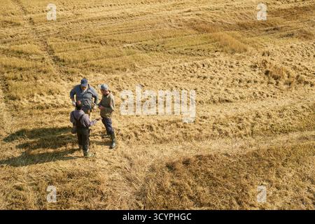 Drei Männer, die auf dem Feld stehen, diskutieren über die Arbeit, tragen Outdoor-Kleidung und -Stiefel, im Kreis positioniert, trockenes Gras und Stoppeln sichtbar herum Stockfoto
