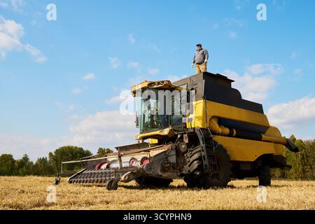 Kaukasischer Mann mittleren Alters, der während der Erntesaison auf einem großen landwirtschaftlichen Mähdrescher auf offenem Feld steht, Ackerland beobachtet und den Maschinenbetrieb überwacht Stockfoto