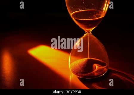 Eine Sanduhr wird dramatisch durch ein warmes, goldenes Licht vor einem dunklen, schwarzen Hintergrund beleuchtet. Stockfoto