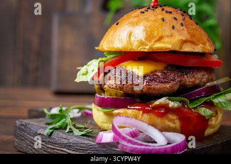 Nahaufnahme von Rinderhamburger mit Tomaten, Käse, eingelegte Gurke, rote Zwiebeln, Salat, Ketchup und Käse, auf Holzbrett. Stockfoto