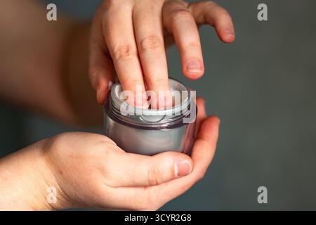 Nahaufnahme der Hand, die in den Gesichtscremebehälter taucht. Detaillierte Aufnahme, die Finger zeigt, wie sie weiße Creme aus einem offenen Behälter sammeln. Premium-Hautpflegeprodukt in dai Stockfoto