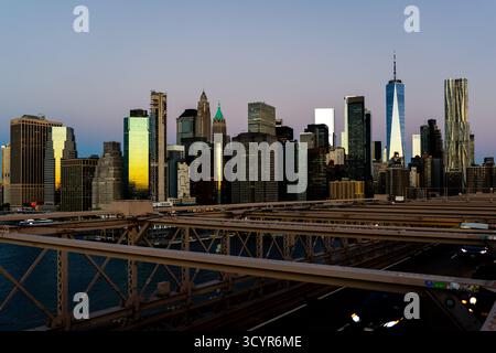 Die Skyline von Manhattan leuchtet bei Sonnenaufgang von der Brooklyn Bridge aus mit Verkehr und dem beleuchteten One World Trade Center. Stockfoto