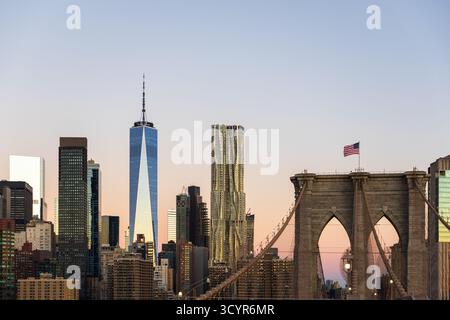 Die Skyline von Lower Manhattan, einschließlich One World Trade Center, steht bei Sonnenaufgang hinter einem Steinturm der Brooklyn Bridge Stockfoto