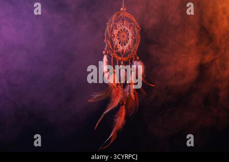 Wunderschöner Traumfänger, der in farbigen Lichtern auf dunklem Hintergrund mit Rauch hängt Stockfoto