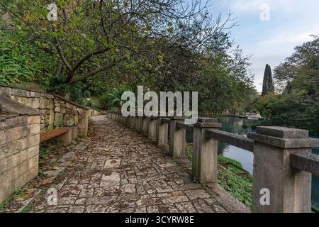 Der malerische Weg ist mit Kopfsteinpflaster umgeben von Steinmauern, umgeben von lebhaftem Grün und Bäumen, die zu einem ruhigen Wasserkörper, Kreatin, führen Stockfoto