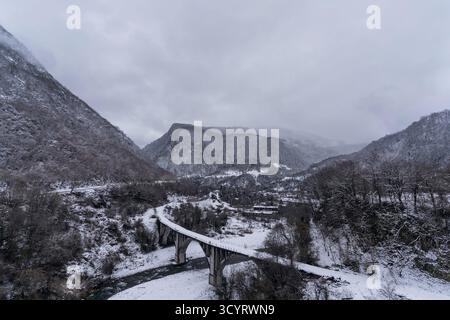 Die schneebedeckte Brücke überspannt anmutig einen sich windenden Fluss in einer ruhigen Berglandschaft, umgeben von Bäumen und Hügeln, die eine ruhige Winterkulisse schafft Stockfoto