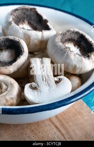 Nahaufnahme von frischen weißen Pilzen in einer Schüssel aus weißer Emaille mit blauem Rand, die auf eine Holzfläche gelegt wird. Stockfoto