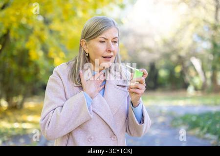 Ältere grauhaarige Frau, die an Halsschmerzen leidet und Sprühmedikamente zur Schmerzlinderung einsetzt. Frau, die im Park läuft. Stockfoto