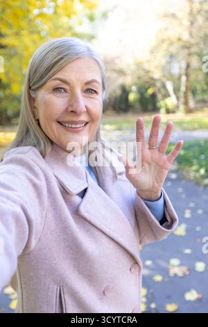 Ältere Frau in einem stilvollen Mantel, die ein Selfie festnimmt, während sie lächelt und der Kamera zuwinkt und den wunderschönen Herbsttag im Park mit goldenem Laub genießt Stockfoto