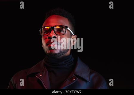 Taille-up Porträt des jungen Schwarzen Mannes in stilvoller Lederjacke und Brille, die auf dunklem Hintergrund in die Ferne schaut Stockfoto