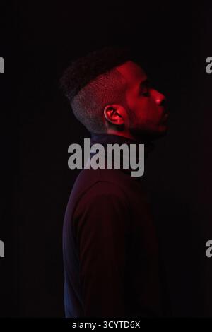 Profilporträt eines jungen gutaussehenden afroamerikanischen Mannes mit stilvollem Haarschnitt, der im Dunkeln mit geschlossenen Augen steht Stockfoto