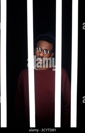 Konzeptuelles Porträt des jungen modischen Schwarzen Mannes in Brille, der hinter Lampen im Dunkeln steht und in die Kamera blickt Stockfoto