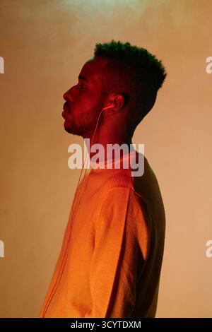 Profilporträt des jungen, stylischen Black Guy mit Ohrhörern, die stehen und in die Ferne schauen, während sie Musik hören Stockfoto