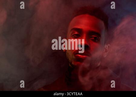 Porträt eines attraktiven Afroamerikaners, umgeben von Rauch, der im Dunkeln steht und mit traurigem Gesichtsausdruck beiseite blickt Stockfoto