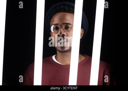 Porträt des hübschen, modischen Schwarzen Mannes, der hinter Leuchtlampen steht und in die Kamera blickt Stockfoto