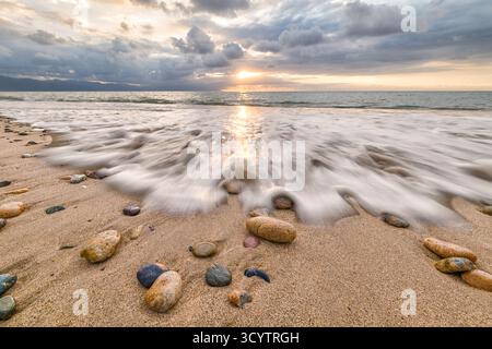 Eine Nahaufnahme des Wassers, das über Felsen fließt, mit Sonnenstrahlen, die durch Wolken am Horizont des Ozeans brechen Stockfoto