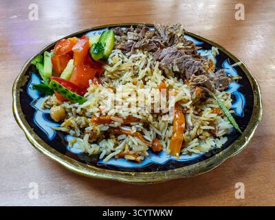 Authentische usbekische Plov (Pilaf) serviert auf einem traditionellen handbemalten Teller mit duftendem Reis, zartem geriebenem Fleisch und einer lebendigen frischen Tomate und Stockfoto