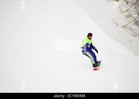 Ein bärtiger Mann in lebendiger Oberbekleidung genießt Snowboarden auf einer verschneiten Piste und bietet Wintersportarten an. Stockfoto