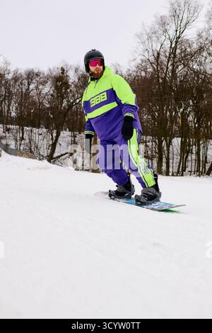 Ein bärtiger Mann genießt im Winter Snowboarden auf einer verschneiten Piste, die von Bäumen umgeben ist. Stockfoto