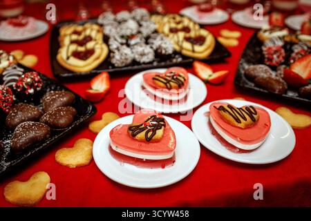 Mehrschichtige Mousse-Desserts in Herzform mit rosafarbener Glasur und Schokoladennebel. Serviert auf weißen Tellern über einem roten Tuch, perfekt für Romantik oder Urlaub Stockfoto