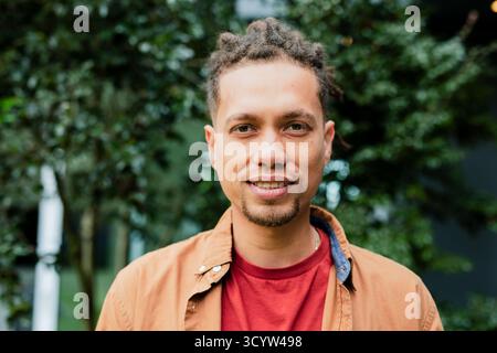 Afroamerikaner, der vor grünem Laub im Garten steht und ein rotes T-Shirt und eine braune Jacke trägt Stockfoto