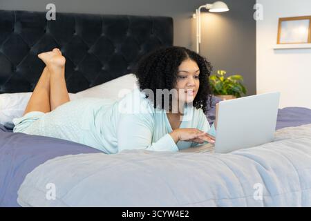 Mittelerwachsene afroamerikanische Frau, die im Schlafzimmer gegen getuftete Kopfteile liegt und auf silbernem Laptop schreibt Stockfoto