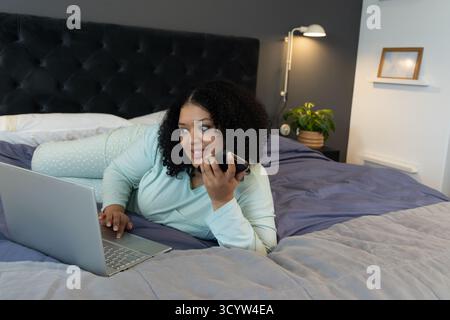 Afroamerikanische Frau, die auf dem Bett liegt und Ellbogen stützt, indem sie im Schlafzimmer per Smartphone auf einem Laptop schreibt Stockfoto