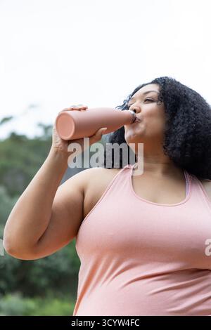Afroamerikanische Frau trinkt aus matten pfirsichfarbenen wiederverwendbaren Wasserflaschen draußen im Park Stockfoto