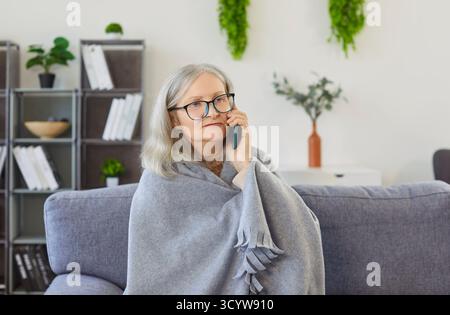 Ernsthafte Seniorin in Decke gewickelt, auf dem Sofa sitzend, mit dem Handy gesprochen und unwohl gefühlt. Stockfoto