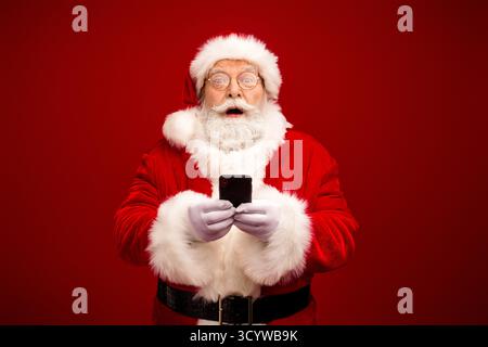 Der Weihnachtsmann in einem roten Anzug mit weißem Pelzbesatz hält ein Smartphone vor einem roten Hintergrund und ist bereit für Weihnachtseinkäufe Stockfoto
