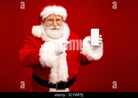 Der Weihnachtsmann zeigt einen leeren Smartphone-Bildschirm und zeigt auf ihn vor einem hellroten Hintergrund für ein Weihnachtsfoto Stockfoto