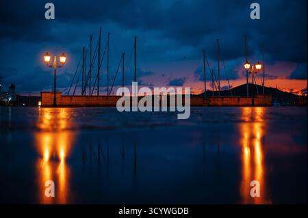Eine ruhige Uferszene in der Abenddämmerung mit Silhouetten von Segelbooten vor einem farbenfrohen Sonnenuntergangshimmel, mit Reflexen auf nassem Pflaster und glühender Straße Stockfoto