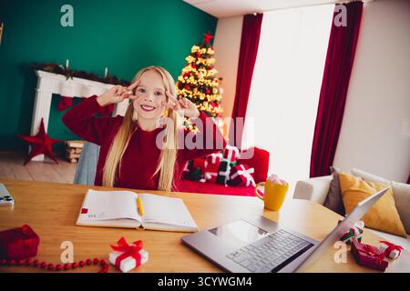 Lustige festliche Mädchen lernen zu Hause mit Weihnachtsbaum-Laptop und Geschenke, die in die Kamera lächeln in einem gemütlichen Wohnzimmer Stockfoto