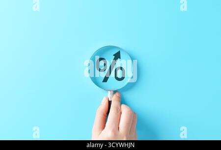 Zinswachstumskonzept mit Vergrößerungsglas, das auf das Prozentsymbol mit einem Aufwärtspfeil auf blauem Hintergrund ausgerichtet ist, das Finanzwesen symbolisiert. Stockfoto
