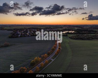 Das Dorf liegt in der Abenddämmerung zwischen Feldern und Bäumen, mit einer gewundenen Straße, die von herbstfarbenen Bäumen gesäumt ist und unter einem glühenden Sonnenuntergang in den Horizont führt Stockfoto