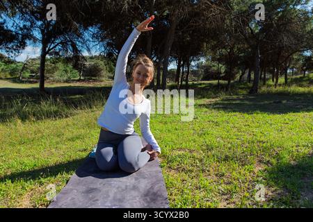 Frau mit weißem Langarmhemd und grauer Leggings kniet auf einer Yoga-Matte in einem sonnigen grasbewachsenen Park, lächelt und macht einen seitlichen Stretch mit einer Stockfoto