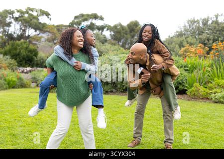 Afroamerikanische Familie lächelt, während sie Kinder mit der Sonnenbrille ihres Vaters auf dem Rücken im Garten trägt Stockfoto