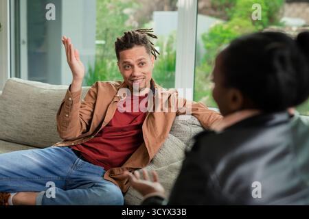 Verschiedene Freunde sitzen auf Sofa mit Kissen im Wohnzimmer und unterhalten sich beim Gesten in der Nähe von Fenstern Stockfoto
